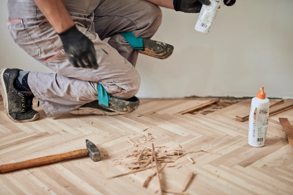 Main ouvrier appliquant un produit de finition sur planches de bois brut avec pinceau plat, portant gants de protection blancs